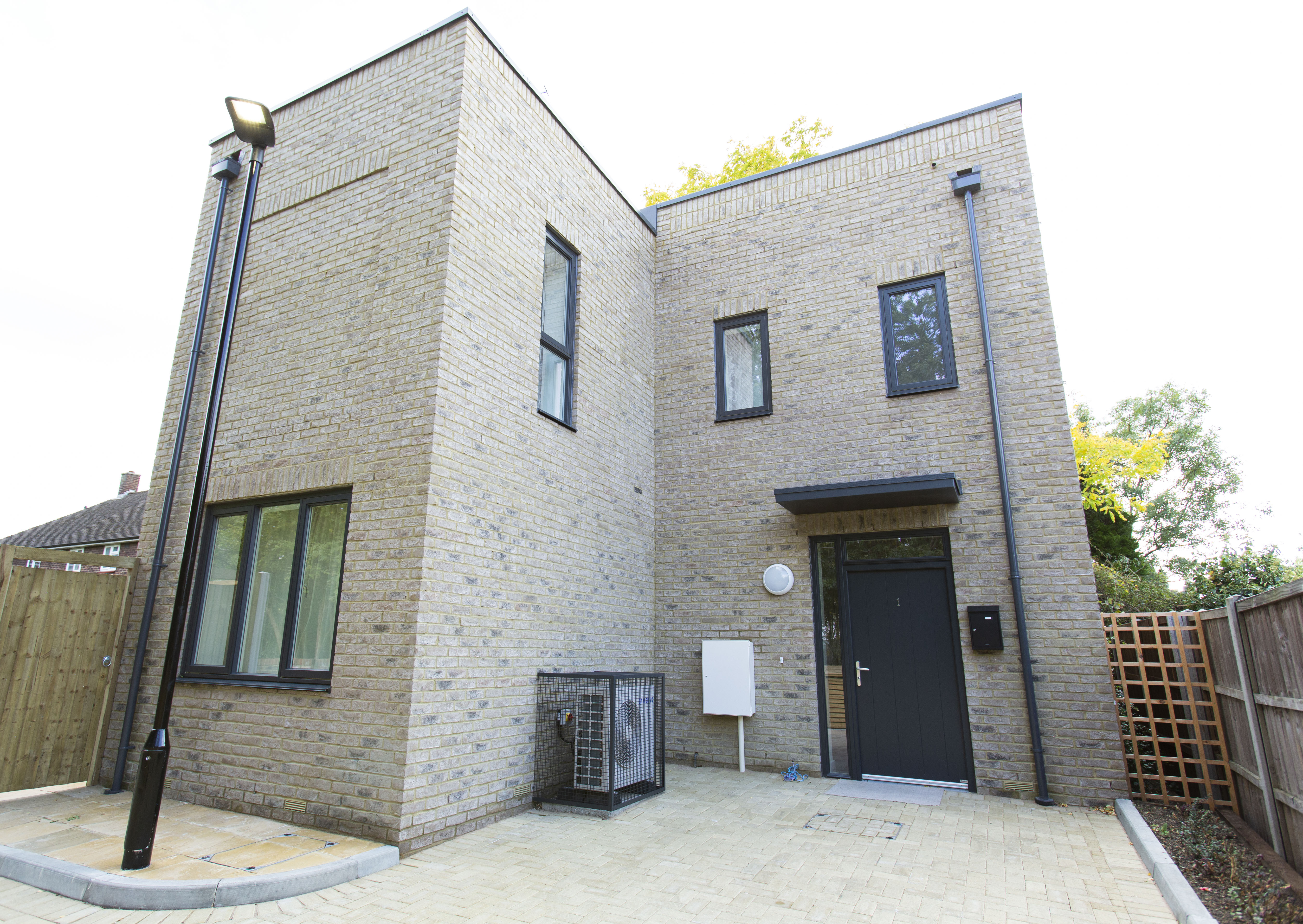 A photograph of a two-storey light brick home with a modern design. A heat pump unit fitted to the house is visible 