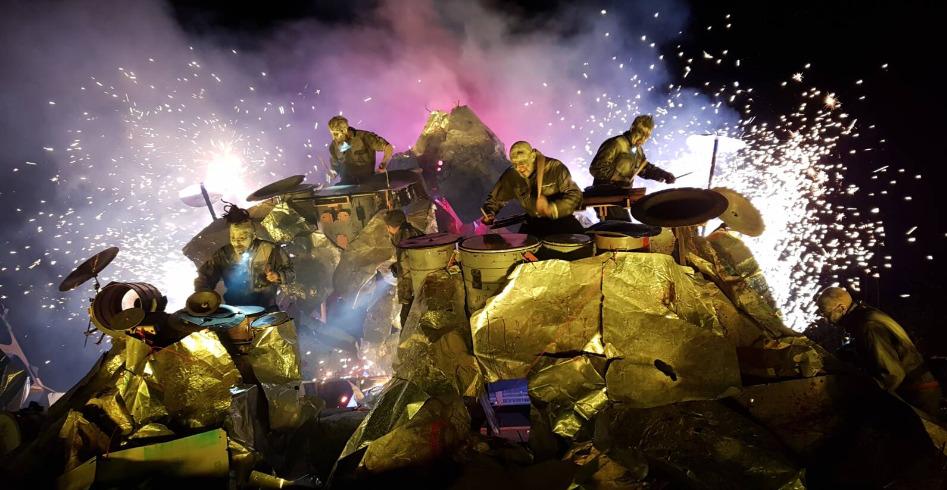 A group of performers dressed in metallic, reflective outfits are seen on a stage that resembles a rocky outcrop. They are engaged in playing various percussion instruments, with dramatic lighting and smoke effects creating a dynamic and intense atmosphere. Bright flashes of light and sparks enhance the visual impact of the performance.