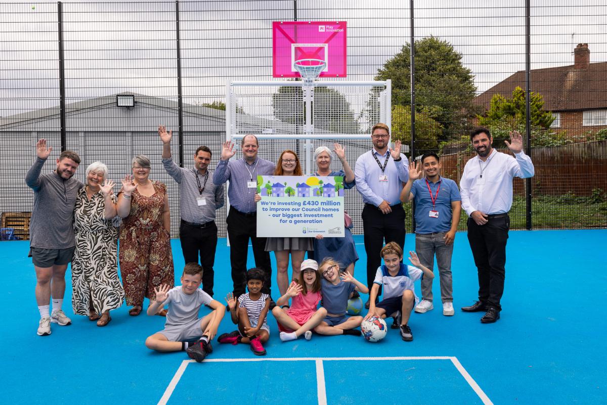 A group consisting of councillors, council staff, centre users and contractors waving in the MUGUA at Middle Park Community Centre logo.  The writing on the board reads: "We're investing £430m to improve council homes - our biggest investment for a generation." 