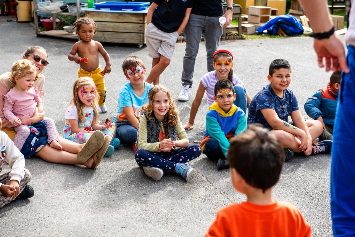 Photo shows group of children celebrating the opening of a new Family Hub in the Royal Borough of Greenwich. Children are sat outside on the ground listening to a story.