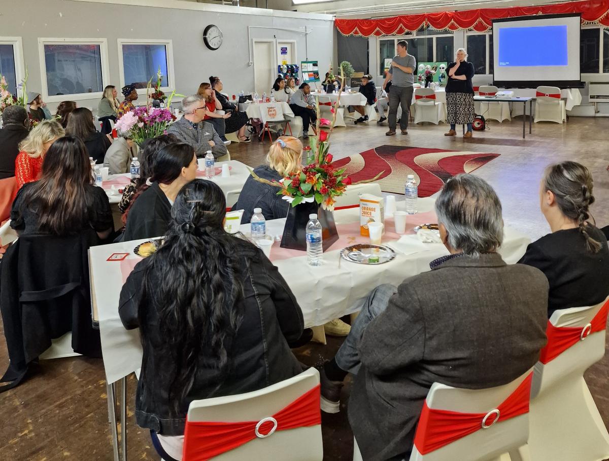 A photo of the inside the C2K Community Centre during the event, people sitting at tables looking to the front where two people stand talking.