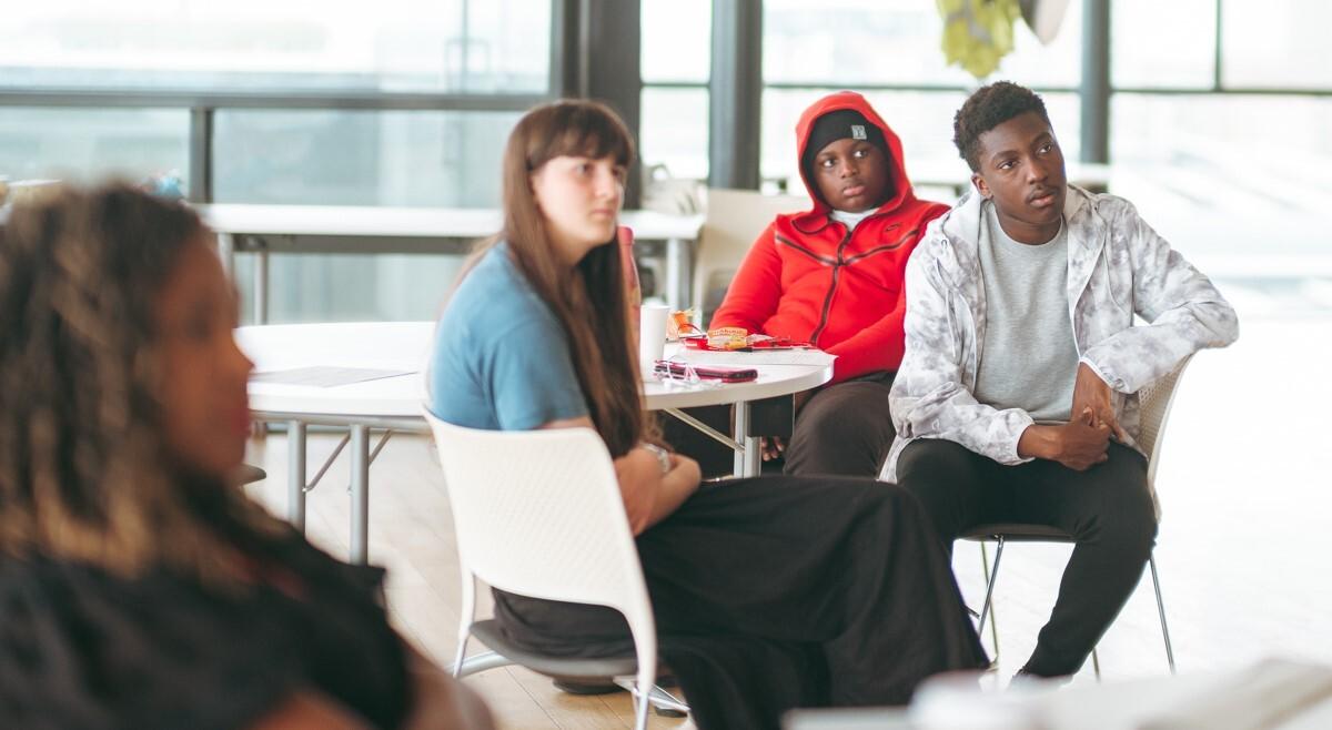 Four young people sit around tables, looking to the front of the room.