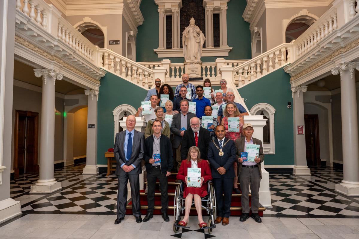 A group of people standing on the steps inside the town hall, all holding up printed certificates