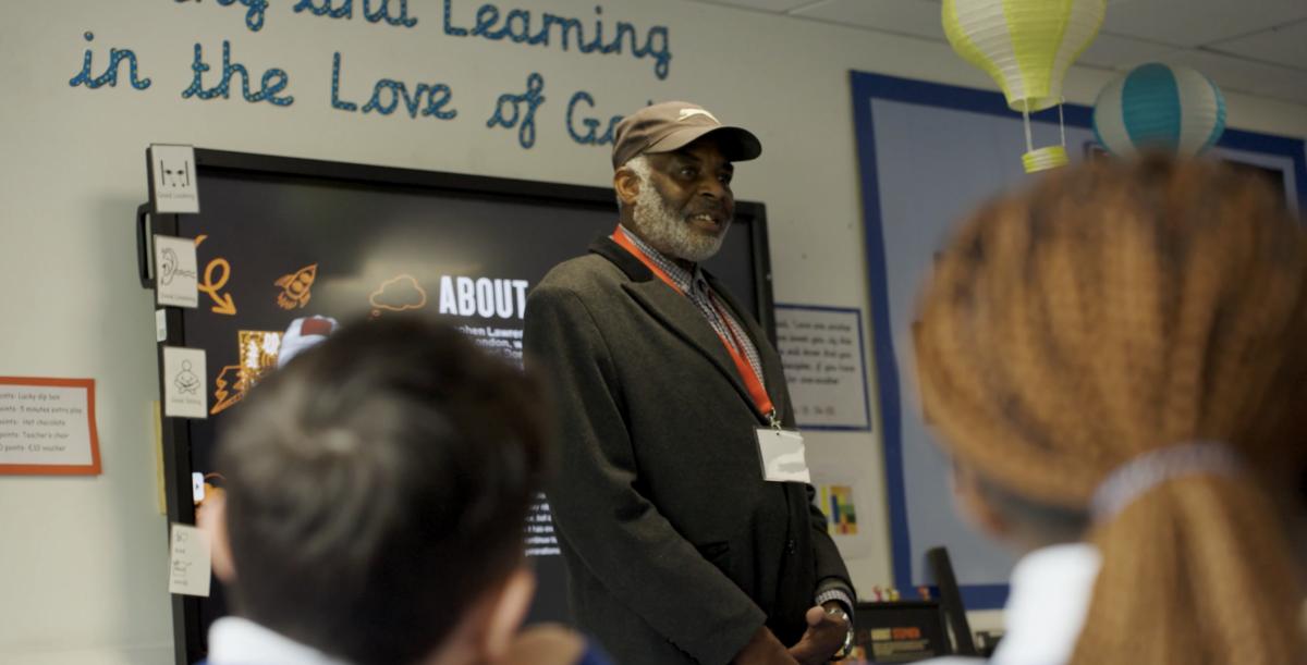 A still from the film, Neville Lawrence standing at the front of a class room with children in the foreground.