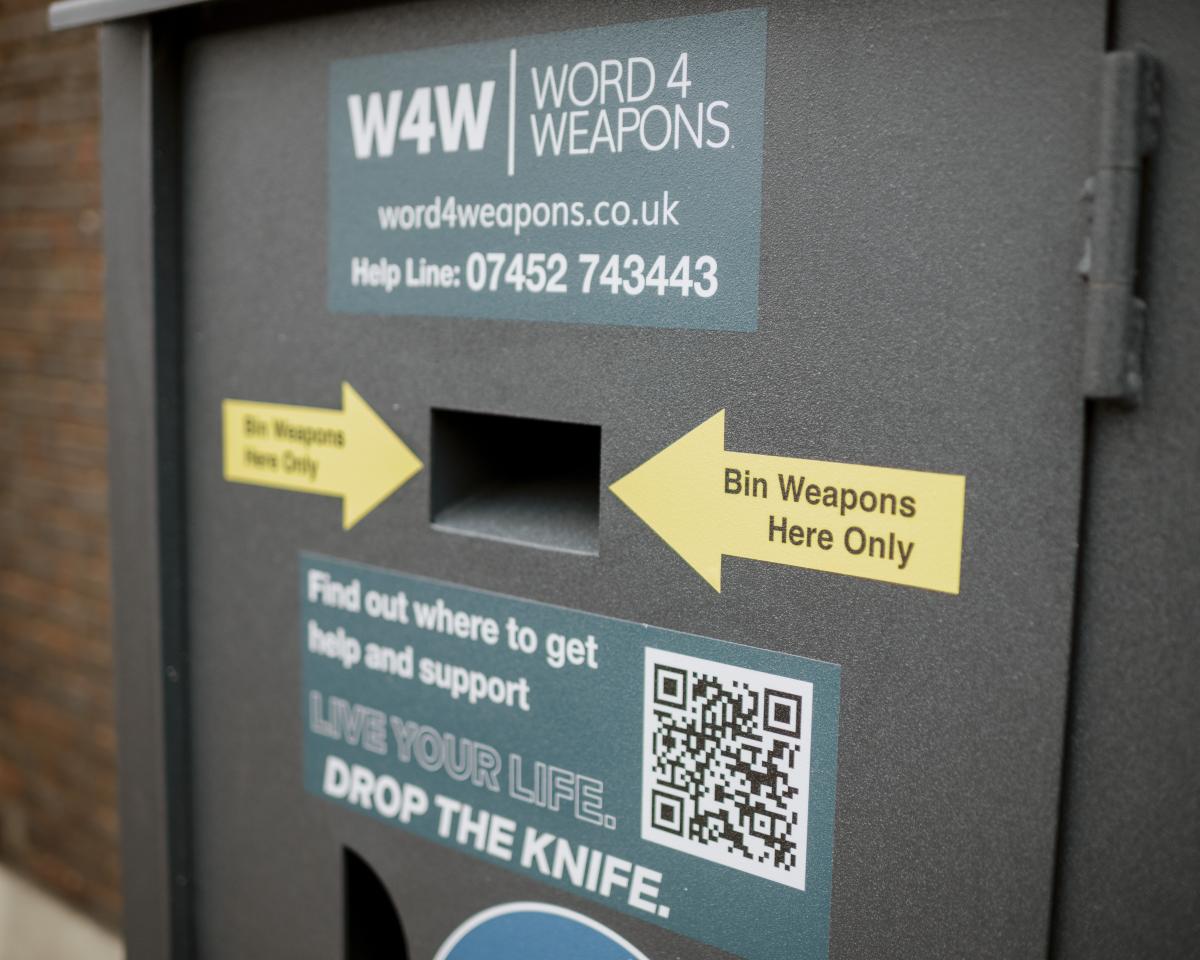 Image of a knife amnesty bin with two yellow arrows pointing to the opening of the bin. Sticker on the bin reading find out where to get help and support. Live your life, drop the knife' and a QR code.