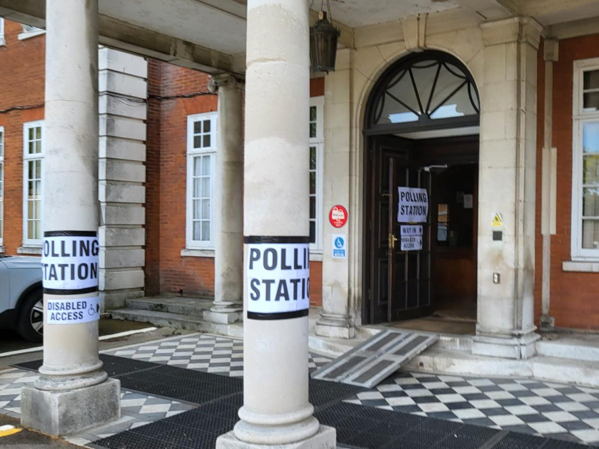 Entrance to a hall with pillars outside. Posters taped on to the pillars reading 'POLLING STATION' and a ramp into the entrance.