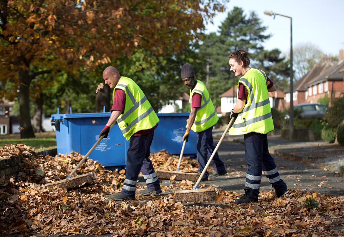 Three people in red council t shirts and high vis vests sweep autumn leaves from the pavement