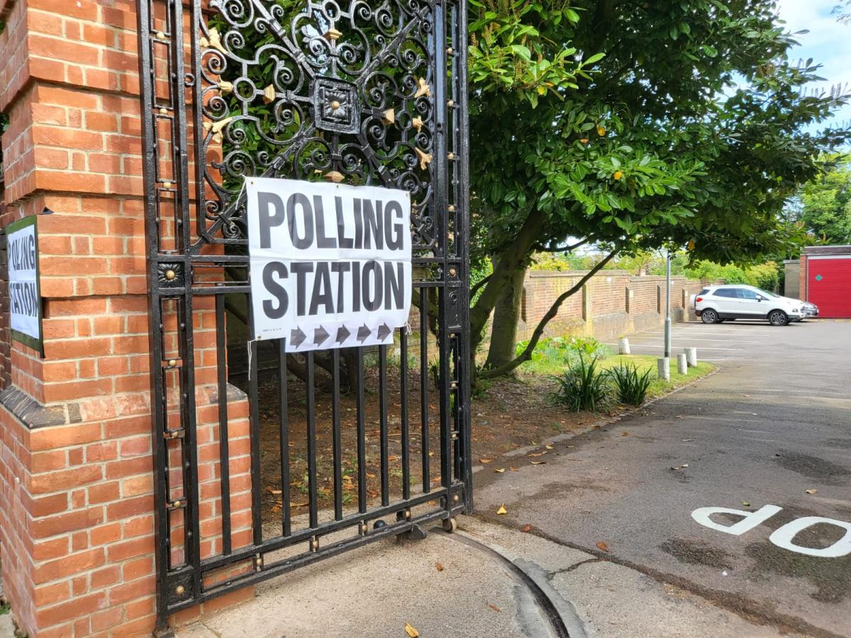 Image of a gate opening in towards a park. A white poster with text reading 'POLLING STATION' fixed on to the gate.