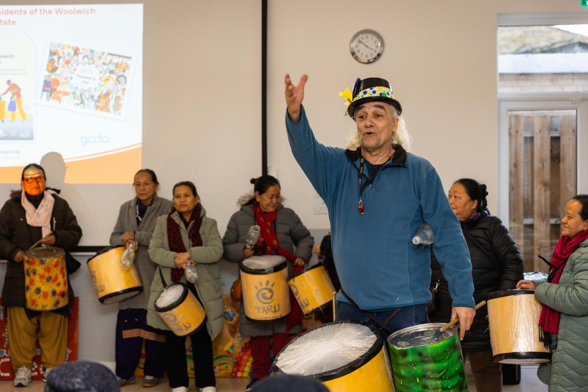 A man is standing with his arm outstretched with a group of women performing a drumming routine.