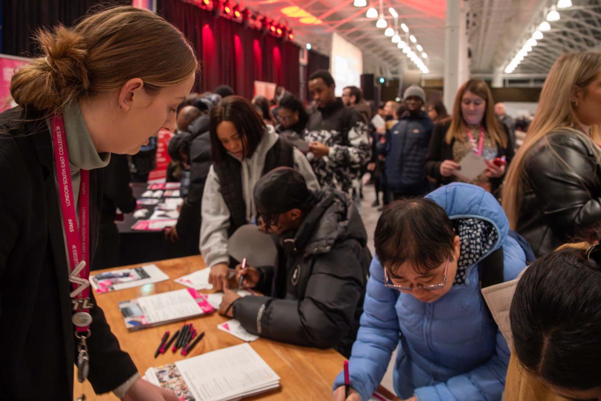 Photo of the summit last year. Woman in a black suit with a red lanyard is stood by a desk talking to another woman in a blue coat who reading information on the desk. Next to her is a teenage boy in a black coat sat down filling in a form