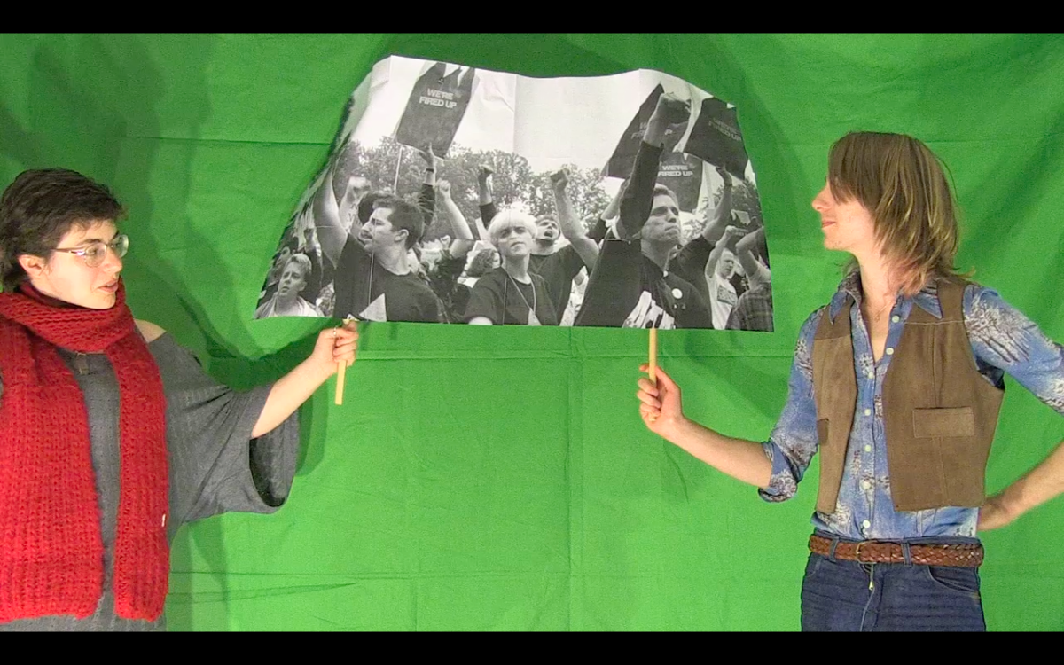 Two people standing facing each other in front of a green screen, holding up a black and white photo of a protest march