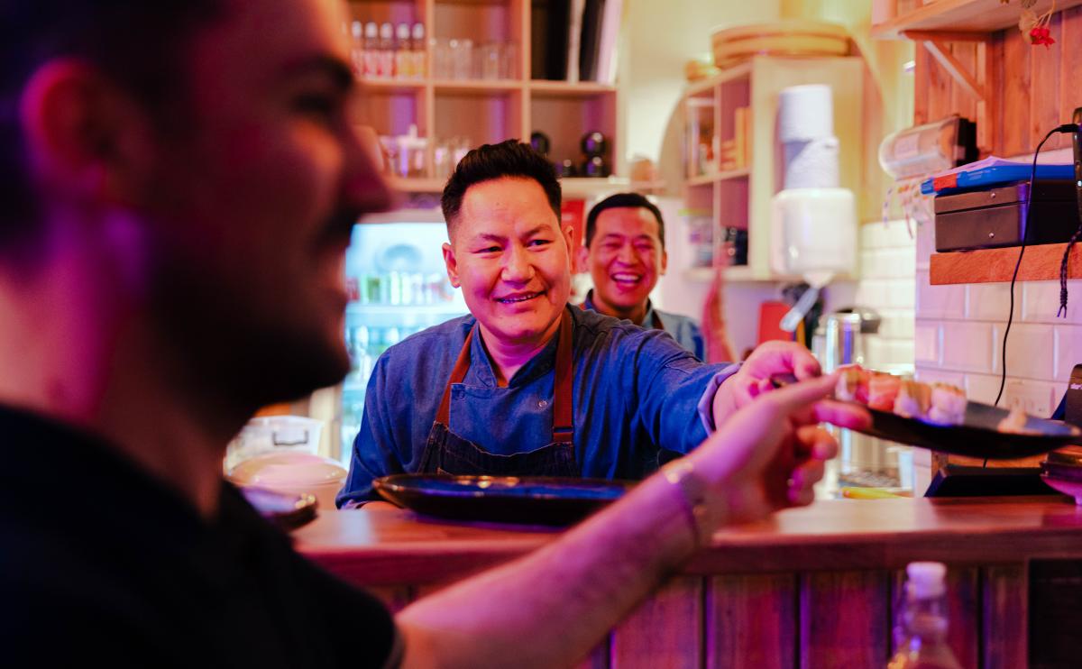 A happy sushi chef in a busy restaurant passing a plate to a waiter.