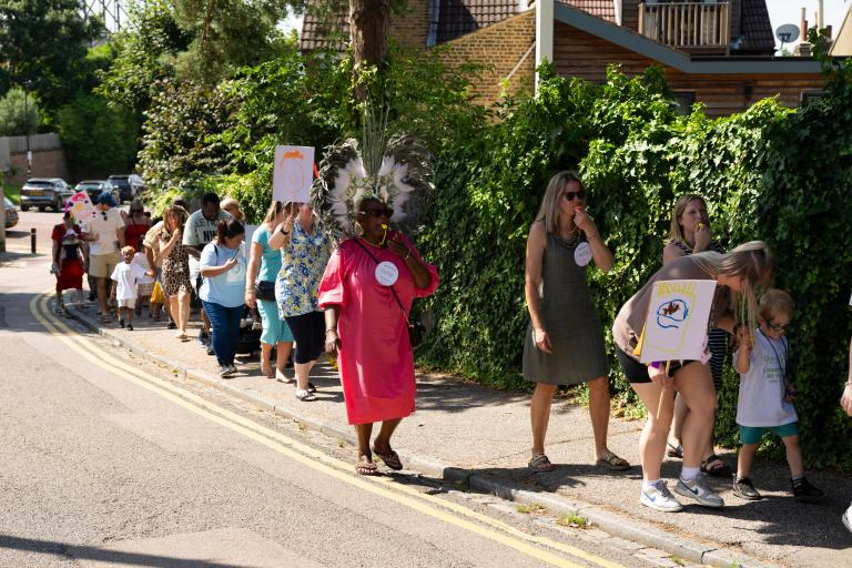 Image shows a parade of adults and children with handmade signs and banners, some adults have whistles, one woman is wearing a big headress.