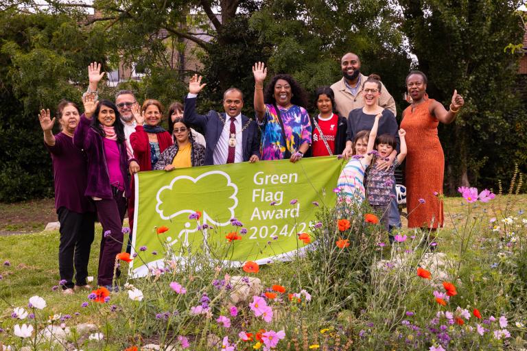 Councillors Jit Ranabhat, Sandra Thomas, Adel Khaireh, and Ann-Marie Cousins with Friends of Plumstead Gardens group members. They are holding a green flag reading "Green Flag Awards 2024/45" and posing behind a bed of wildflowers.
