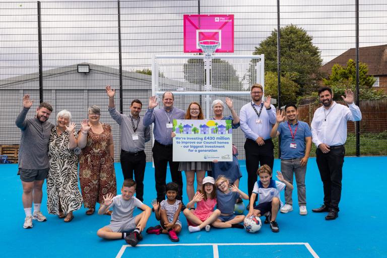 A group consisting of councillors, council staff, centre users and contractors waving in the MUGUA at Middle Park Community Centre logo.  The writing on the board reads: "We're investing £430m to improve council homes - our biggest investment for a generation." 