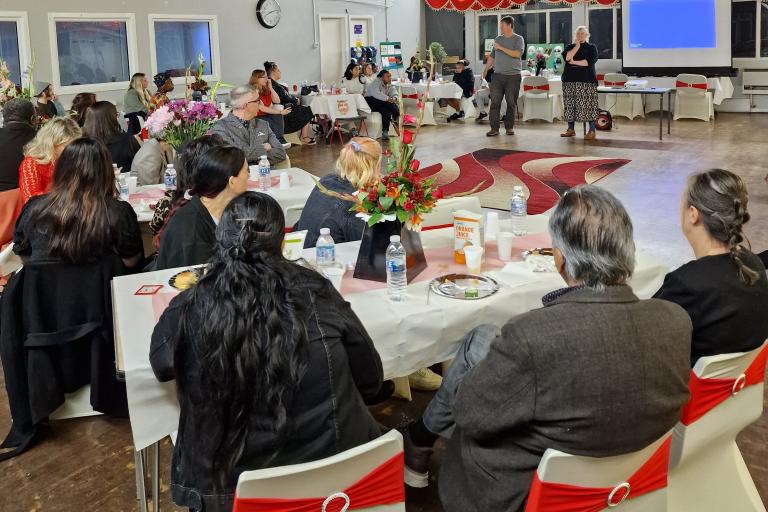 A photo of the inside the C2K Community Centre during the event, people sitting at tables looking to the front where two people stand talking.