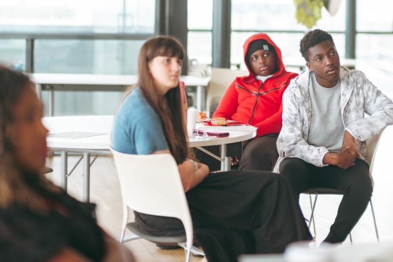 Four young people sit around tables, looking to the front of the room.