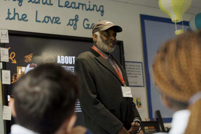 A still from the film, Neville Lawrence standing at the front of a class room with children in the foreground.