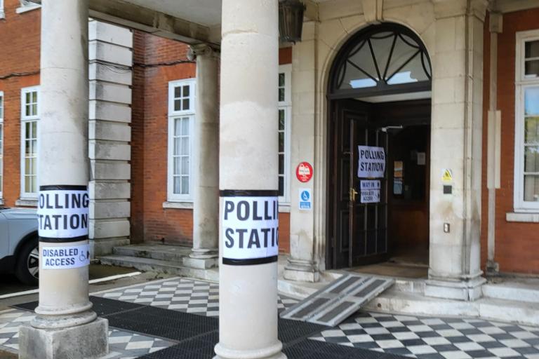 Entrance to a hall with pillars outside. Posters taped on to the pillars reading 'POLLING STATION' and a ramp into the entrance.