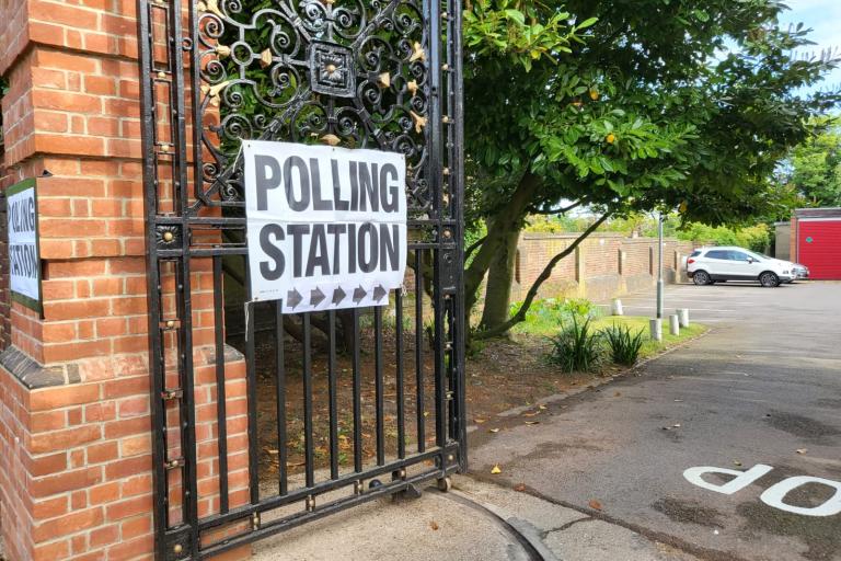 Image of a gate opening in towards a park. A white poster with text reading 'POLLING STATION' fixed on to the gate.