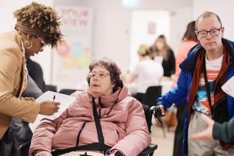 a member of staff filling in a form and talking to a female tenant in a wheelchair next to her partner.