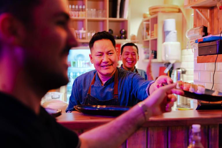 A happy sushi chef in a busy restaurant passing a plate to a waiter.