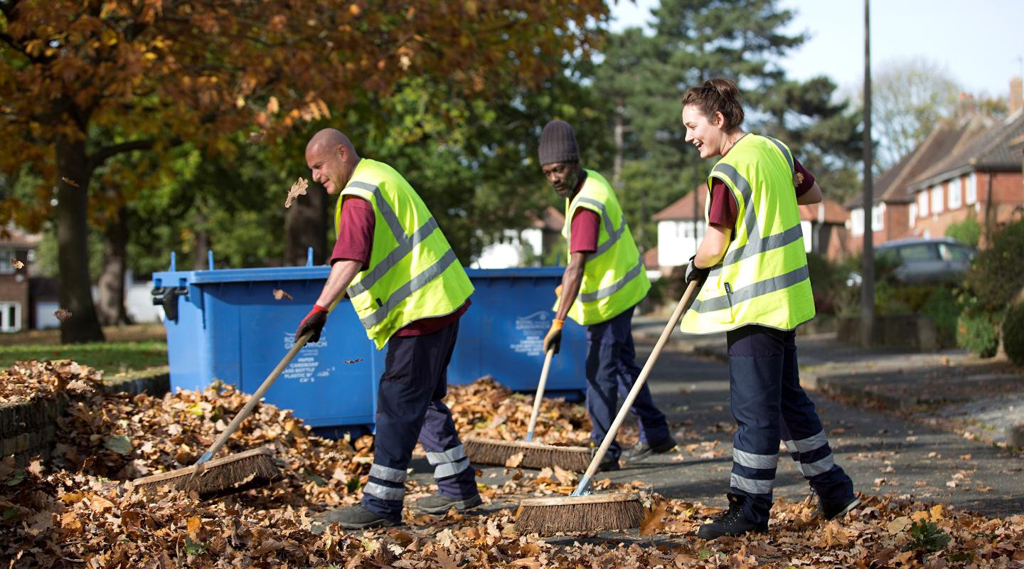 Three people in red council t shirts and high vis vests sweep autumn leaves from the pavement