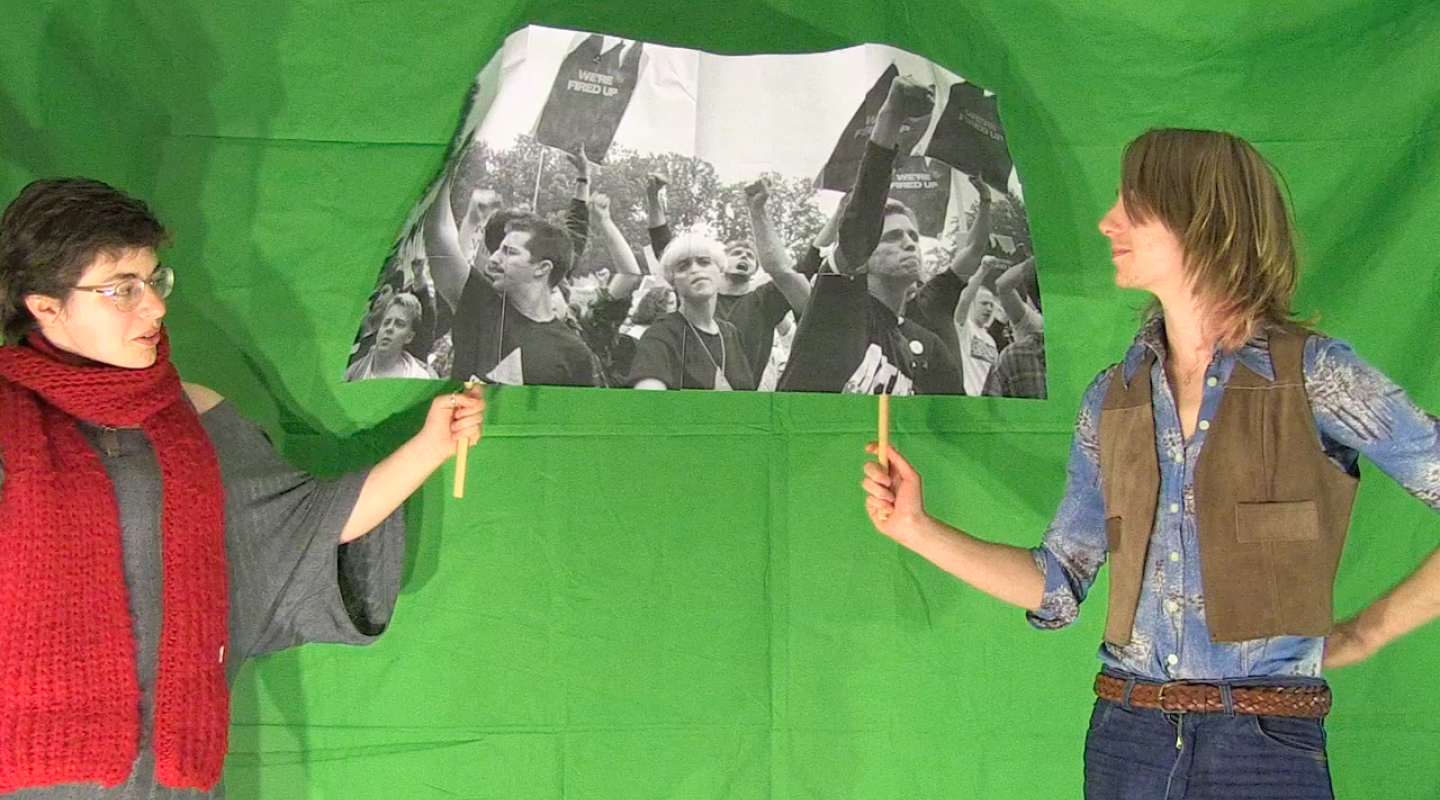 Two people standing facing each other in front of a green screen, holding up a black and white photo of a protest march